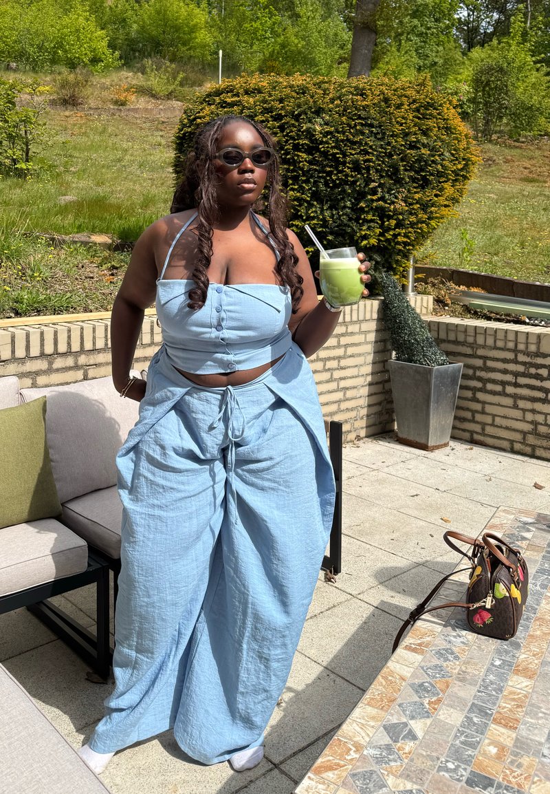 Femme en tenue bleu clair et lunettes de soleil tenant une boisson verte avec une paille, debout sur un patio avec du mobilier d'extérieur et de la verdure.