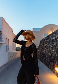 Cardigan in pizzo nero con maniche lunghe, apertura frontale, indossato sopra pantaloni neri. Cappello in paglia e borsa con manico decorativo. Sfondo di muro in pietra.