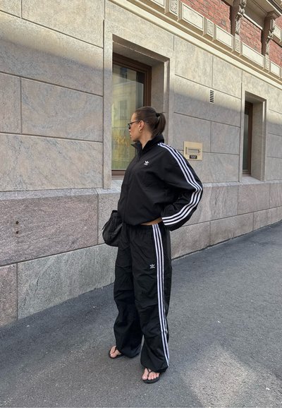 Chaqueta y pantalones de chándal negros con rayas blancas, hechos de un material ligero. El conjunto presenta un diseño deportivo y un cuello con cremallera.