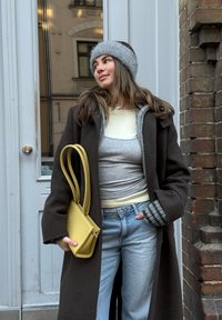 Jeune femme portant un bandeau gris, un manteau brun foncé, des couches de hauts, et un jean clair, tenant un sac à main jaune, debout près d'une porte et d'un mur en briques.