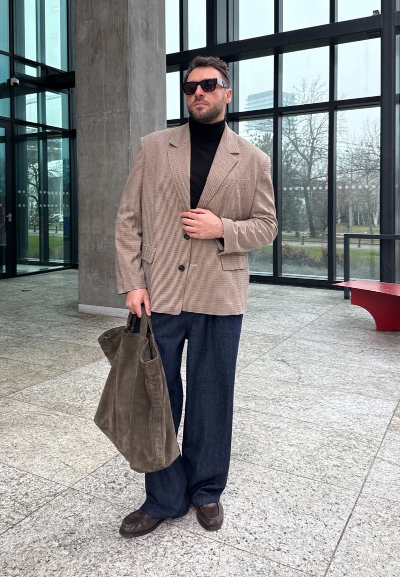 Homme portant un blazer beige, un col roulé noir, un pantalon foncé, des lunettes de soleil, tenant un grand sac en daim marron à l'intérieur d'un bâtiment en verre.