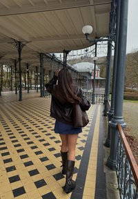 Femme aux cheveux longs portant une veste sombre, une jupe bleue et des bottes noires, marchant sur un sol carrelé à motifs sous un pavillon en fer couvert.