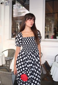 Giovane donna in un abito a pois bianco e nero che tiene in mano una rosa rossa, mentre sta in una terrazza di un caffè con tavoli e sedie nei dintorni.