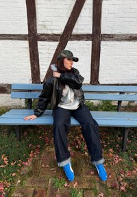 Ung kvinde iført camouflagekasket, sort læderjakke, grå hættetrøje og blå sneakers sidder på en lyseblå bænk mod en hvid murstensvæg.