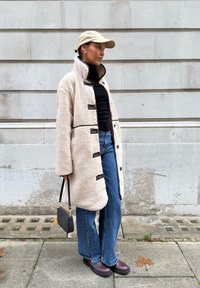 Manteau en peau retournée couleur crème avec des boutons noirs, associé à un col roulé noir, un jean évasé bleu et des bottines marron. Sac à main noir inclus.