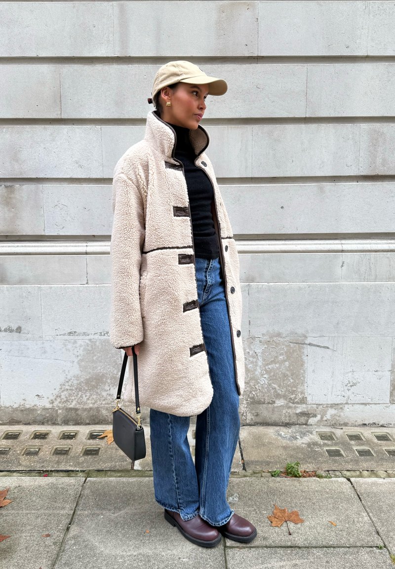 Manteau en peau retournée couleur crème avec des boutons noirs, associé à un col roulé noir, un jean évasé bleu et des bottines marron. Sac à main noir inclus.