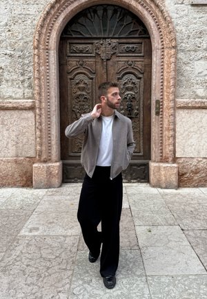 Cárdigan beige con cremallera sobre una camiseta blanca, combinado con pantalones negros de pierna ancha y zapatos negros, de pie cerca de puertas de madera ornamentadas.