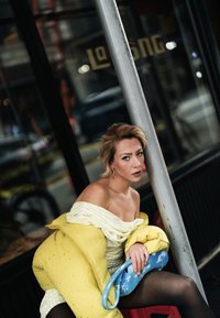 Kollane pufijakk kaunistustega, valge triibuline õlgadeta kleit, sinine käekott pilvemustriga, mustad sukkpüksid, istub posti kõrval.