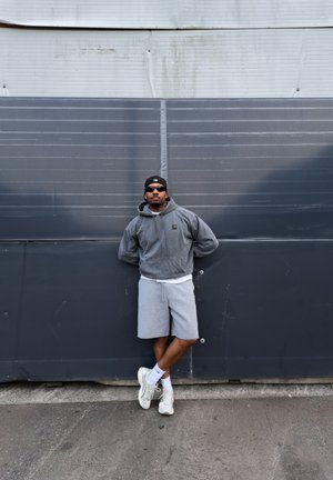 Hombre con sudadera con capucha gris, pantalones cortos grises, zapatillas blancas, gorra negra y gafas de sol apoyado contra una pared industrial gris con las piernas cruzadas.