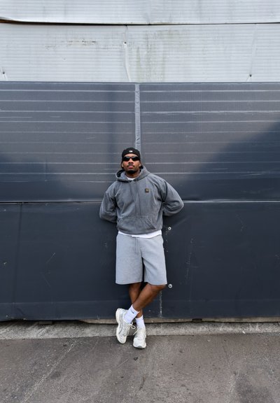 Hombre con sudadera con capucha gris, pantalones cortos grises, zapatillas blancas, gorra negra y gafas de sol apoyado contra una pared industrial gris con las piernas cruzadas.