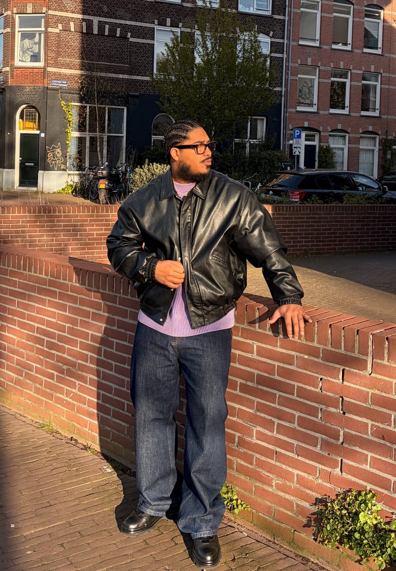 Homme aux cheveux tressés et lunettes, portant une veste en cuir noire et un jean, appuyé contre un mur en briques dans un cadre urbain ensoleillé.
