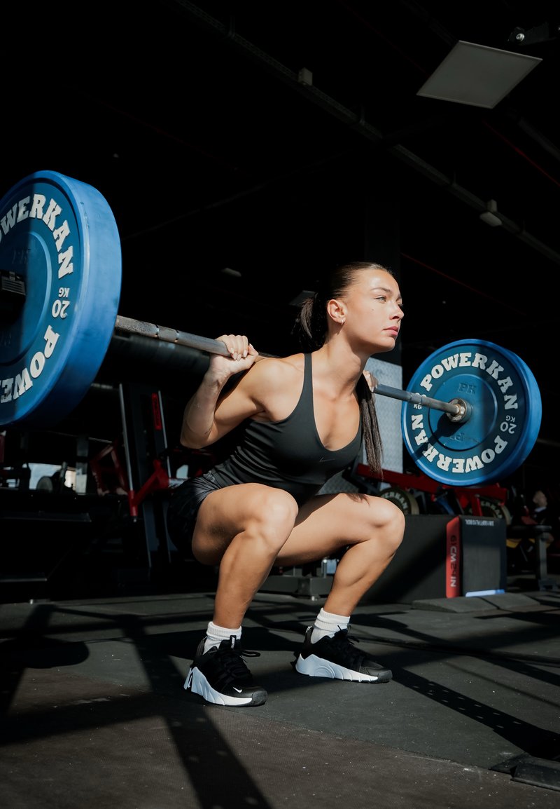 Kvinde i sort tanktop og shorts laver squat med en vægtstang på skuldrene i et fitnesscenter, fokuseret og løfter med korrekt teknik.