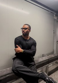 Chemise noire à manches longues côtelée, pantalon noir et chaussures noires. Une montre en or et des lunettes rectangulaires complètent la tenue. Fond lisse et neutre.