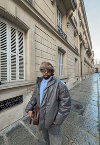 Doudoune grise, chemise bleu clair, lunettes de soleil foncées, chapeau doublé de fourrure et sac bandoulière en cuir marron. Rue pavée et bâtiment classique en arrière-plan.