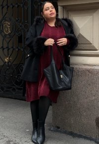 Femme en manteau noir avec des poignets en fourrure et une robe bordeaux se tient contre un mur de pierre, tenant un sac à main en cuir noir.