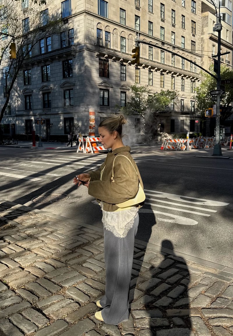 Femme portant une veste marron et un pantalon gris, debout sur un trottoir pavé, regardant son téléphone près de vapeur et de barrières de construction.