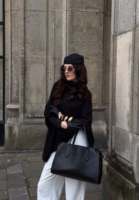 Sac en cuir noir avec deux poignées et une texture lisse, porté avec un poncho noir, un pantalon blanc et des bracelets avec accents dorés.