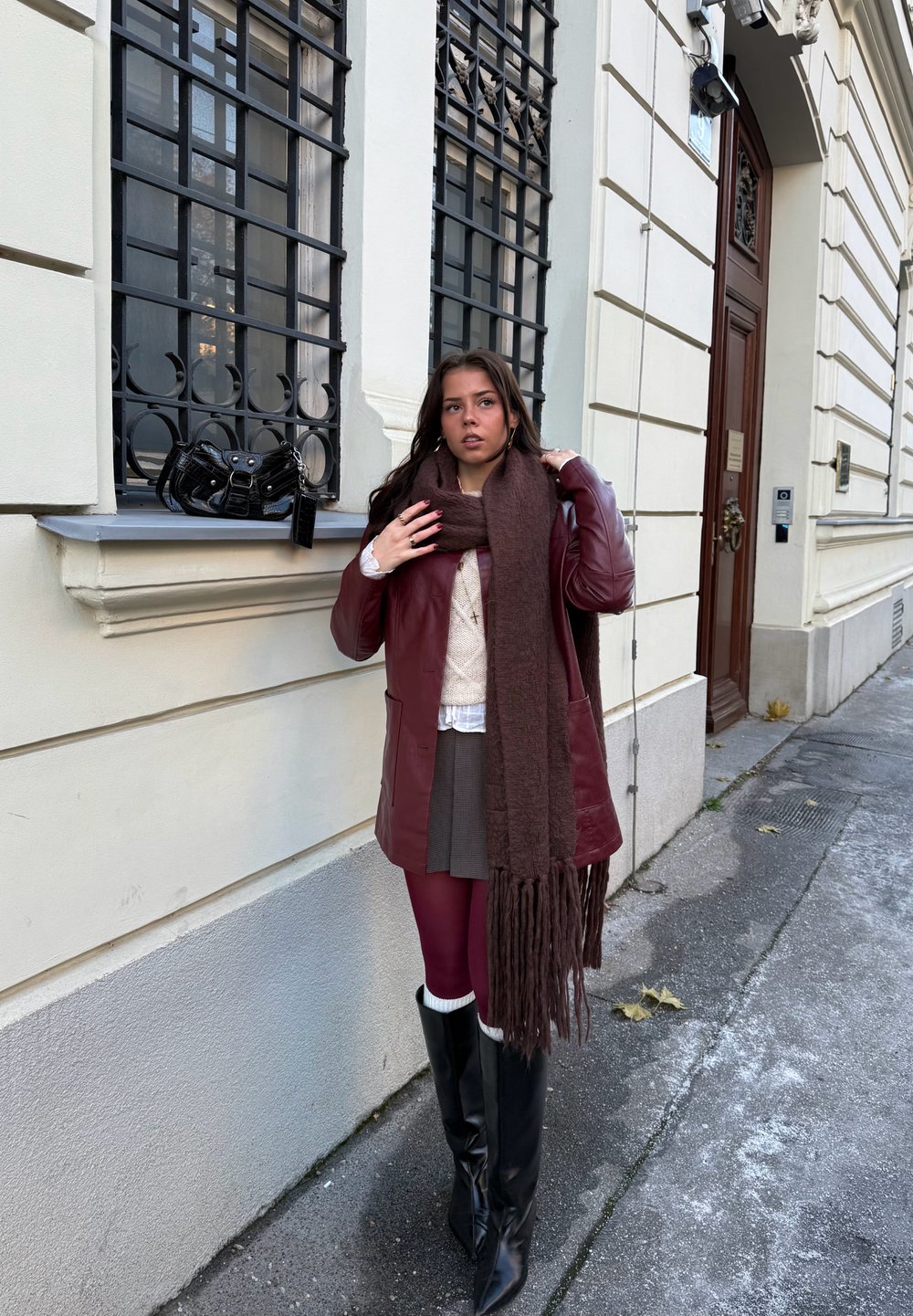 Manteau en cuir marron, pull en tricot blanc, jupe grise, collants bordeaux, bottes hautes noires, écharpe à franges marron ; sac à main noir posé sur un rebord de fenêtre.