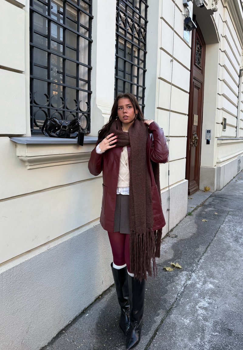 Manteau en cuir marron, pull en tricot blanc, jupe grise, collants bordeaux, bottes hautes noires, écharpe à franges marron ; sac à main noir posé sur un rebord de fenêtre.