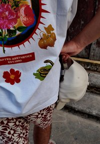 Άτομο που φοράει σορτς με λουλουδάτο μοτίβο και λευκό t-shirt με γραφικά, κρατώντας ένα λευκό καπέλο μπέιζμπολ δίπλα του σε εξωτερικό χώρο.