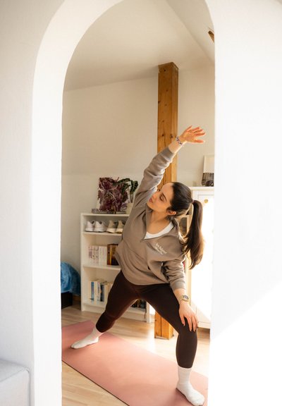 Sudadera gris claro, leggings oscuros y calcetines blancos; realizando una pose de yoga sobre un tapete rosa en una habitación bien iluminada con acentos de madera.