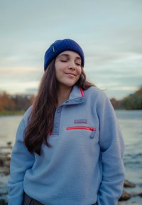 Světle modrý fleecový pulovr s červenou kapsou na zip, šedými doplňky a l�ímcem. Nošený s modrou pletenou čepicí. Pozadí tvoří voda a stromy.