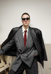 Veste de costume gris croisée, cravate rouge, chemise blanche et manteau en cuir noir. Lunettes de soleil noires et jeans foncés complètent le look.