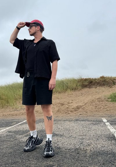 Camisa negra de manga corta sobre una camiseta de tirantes negra, pantalones cortos negros y zapatillas con acentos blancos. Una gorra roja y un tatuaje de mariposa en la pierna.
