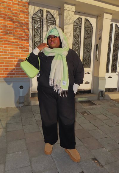 Persona con atuendo negro, bufanda verde claro y crema, gorra de béisbol verde claro y bolso verde a juego, de pie en una acera frente a unas puertas.