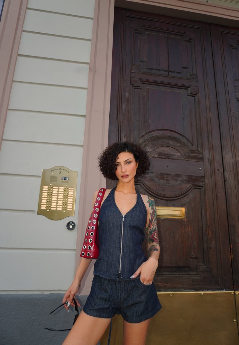 Femme aux cheveux bouclés et tatouage sur le bras, debout devant une grande porte en bois, tenant des lunettes de soleil et portant un sac à épaule rouge et une tenue sombre.