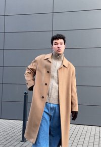 Manteau en laine beige avec des boutons noirs, porté sur un col roulé côtelé beige clair et un jean en denim bleu. Fond gris avec mur texturé.
