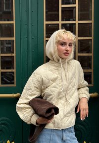 Elfenbeinfarbene steppte Jacke mit Kordelzugkapuze, kombiniert mit lässigen blauen Jeans und einem dunklen braunen Fleece. Grüner gemusterter Hintergrund.