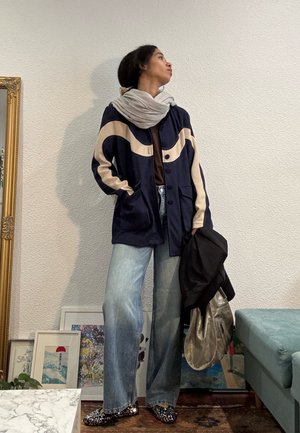 Joven con chaqueta azul marino y beige, bufanda gris, vaqueros anchos azules, sosteniendo bolsas negras y metálicas, de pie junto a una obra de arte enmarcada en el suelo.