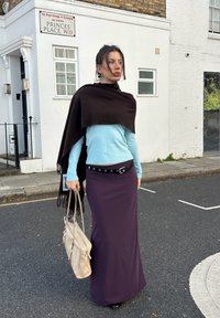 Lang lilla nederdel med et sort bælte, lyseblå langærmet top og en brun sjal. Holder en beige taske. Bygade baggrund.