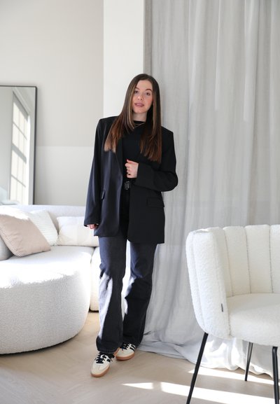 Blazer negro a medida sobre una camisa negra, combinado con jeans de mezclilla oscuros y zapatillas blancas con detalles en negro. Sofá y silla blancos y suaves en el fondo.