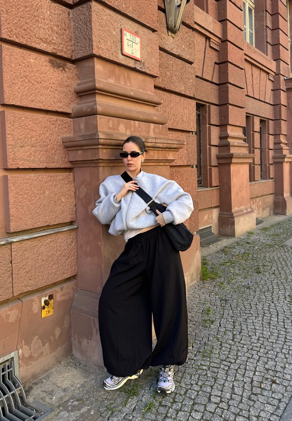 Junge Frau trägt übergroßen grauen Pullover und schwarze weite Hose und lehnt an einem strukturierten Backsteingebäude in einer Kopfsteinpflasterstraße.