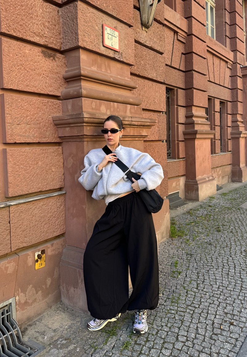 Fiatal nő, vörös színű, túlméretezett szürke pulóvert és fekete bő nadrágot visel, és egy mintás téglafalú épületnek dől macskaköves utcán.