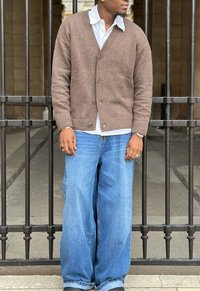 Brown knitted cardigan over a white shirt, teamed with loose blue denim jeans. The scene includes a metal gate in the background.