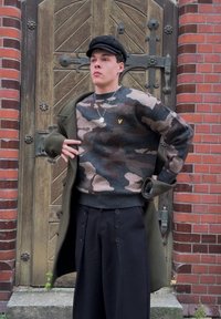 Pletený svetr se vzorem maskáče v béžových a zelených tónech, doplněný širokými černými kalhotami s knoflíkovými detaily, vrstvený s olivově zeleným kabátem.