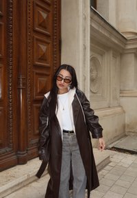 Un long manteau en simili cuir marron sur une chemise blanche, associé à un jean large gris. Des lunettes de soleil noires rectangulaires et des bijoux minimalistes complètent le look.