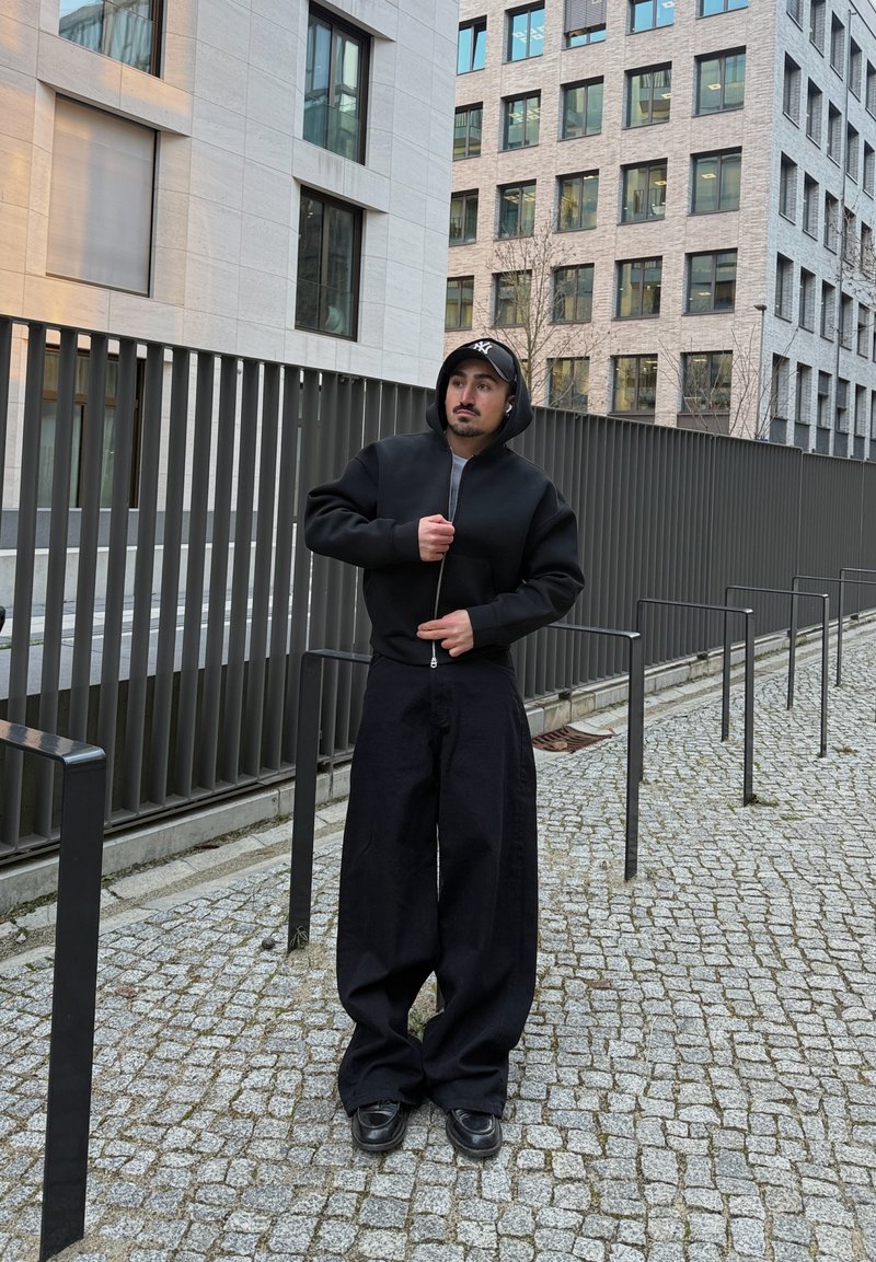 Homme portant un sweat à capuche noir, un pantalon large noir et une casquette noire, debout sur un chemin pavé à côté d'un bâtiment moderne avec des rampes en métal.