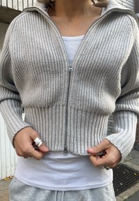 Lysegrå kort genser med ribbet tekstur, glidelås foran og lange ermer. Brukt over en hvit singlet, med flere sølvringer.