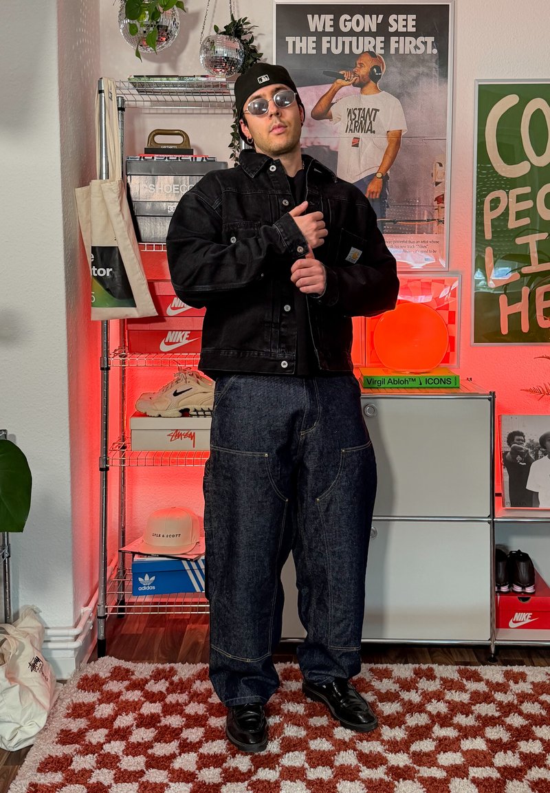 Homme en veste noire, lunettes de soleil et casquette, debout dans une pièce avec des boîtes de baskets, des affiches et un éclairage ambiant rouge.