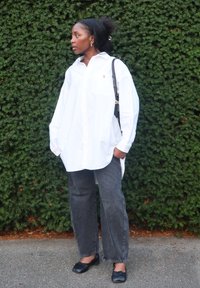 Biała, oversize koszula zapinana na guziki, szare luźne dżinsy, czarne baleriny, z torebką na ramię. Teksturowane zielone tło liściaste.