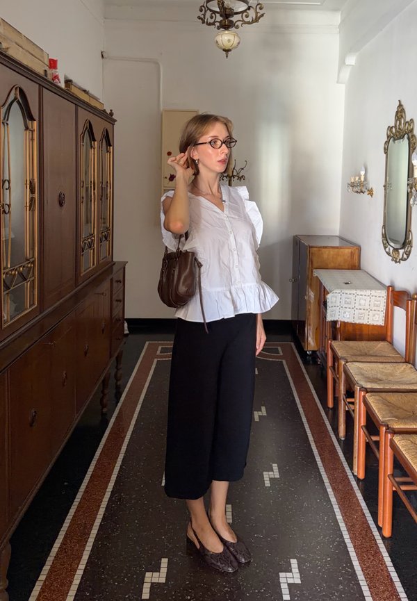 Femme portant des lunettes, un chemisier blanc et un pantalon noir, debout dans une pièce vintage avec des meubles en bois, un miroir et un chandelier.