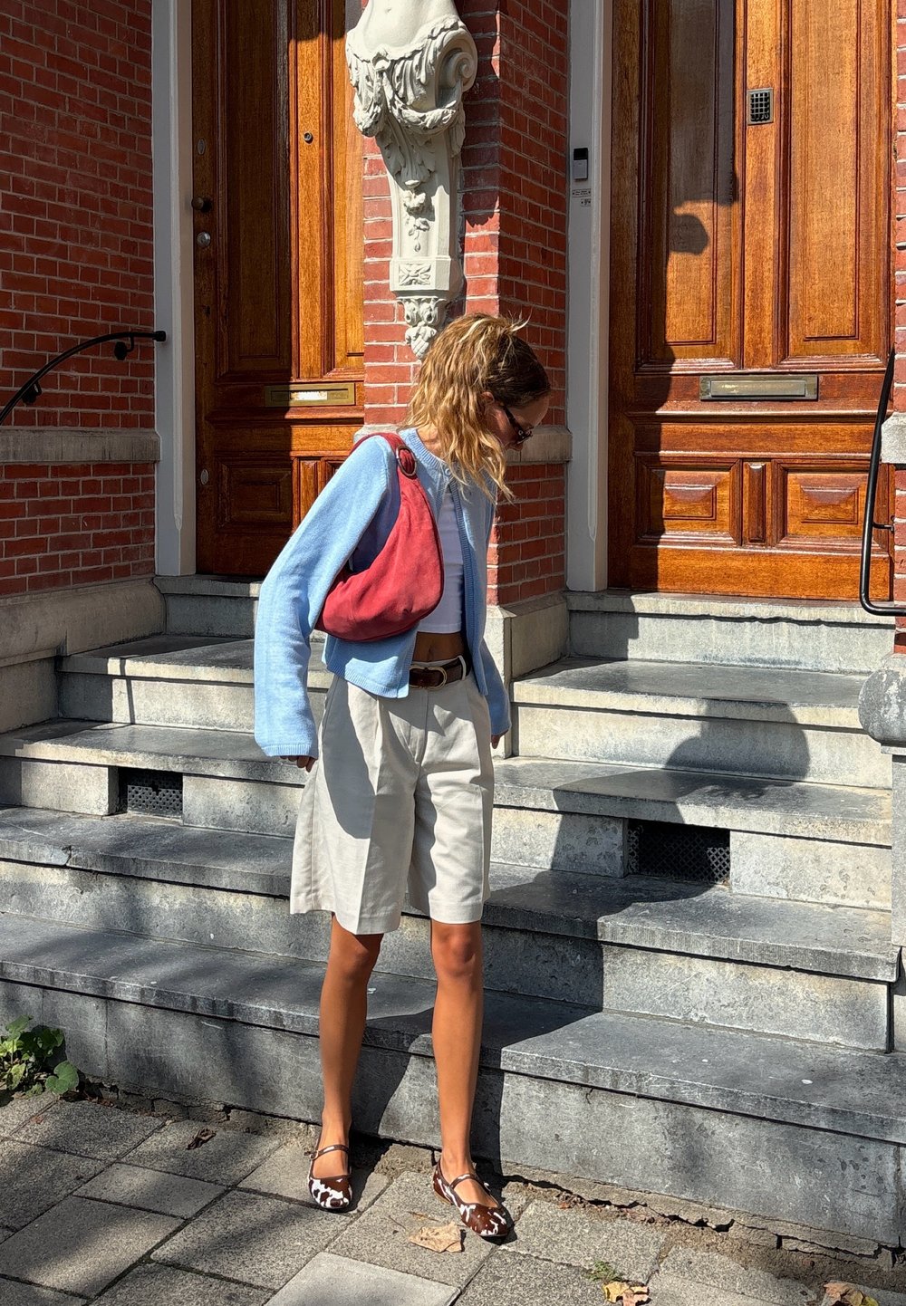 Cardigan azul claro, curto, emparelhado com calções bege. Bolsa vermelha de ombro e sapatos baços castanhos com padrão. Escadas e uma parede de tijolos ao fundo.