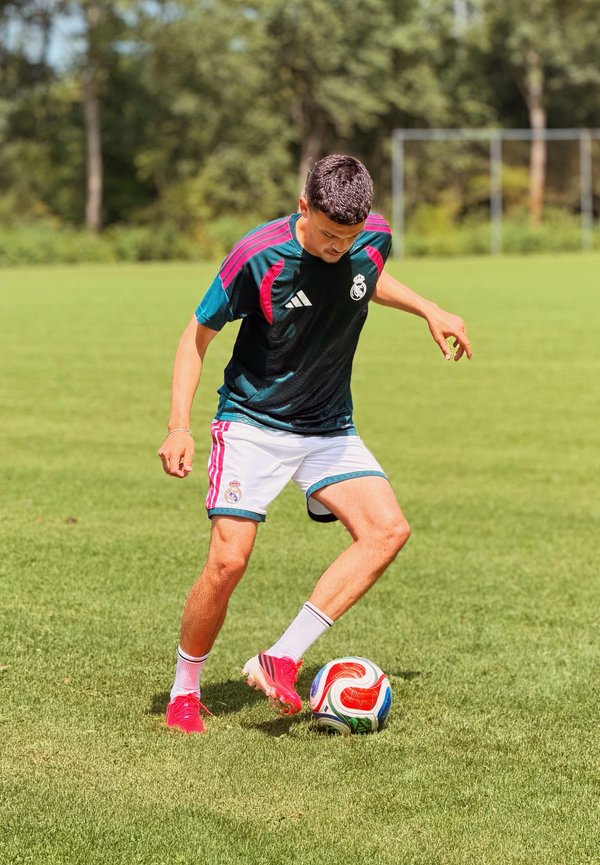 Jeune joueur de football masculin en tenue d'entraînement du Real Madrid contrôlant un ballon coloré sur un terrain herbeux avec des arbres en arrière-plan.
