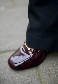 Bordeaux leren loafers met patentafwerking, goudkleurig metalen accent en vierkante neus, gecombineerd met zwarte broeken, rustend op een gestructureerde grijze ondergrond.