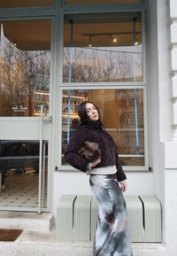 Γυναίκα με σκούρο σακάκι και φούστα με σχέδιο ποζάρει με ένα καφέ τσαντάκι έξω από καφέ με μεγάλα τζάμια και ορατά εσωτερικά ράφια.