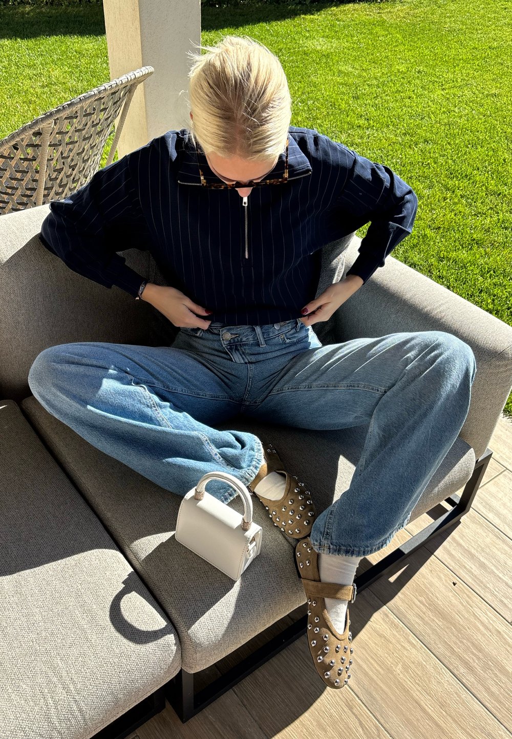 Osoba sedící na venkovním gauči s nohama zkříženýma, upravující námořnický pruhovaný top, má na sobě modré džíny, hnědé boty s cvočky, bílé ponožky a drží bílou kabelku.